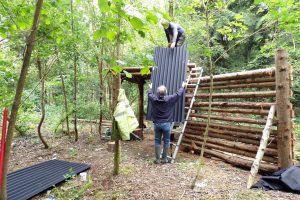 Two volunteers lifting a corrugated waterproof roofing sheet onto a log-built wildlife hide in the woodland.