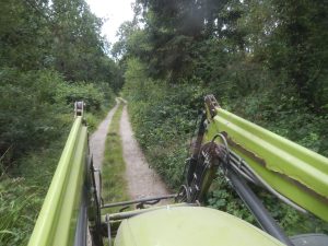 View from a tractor cab looking through the raised loader arms down a winding gravel track through Howes Wood, surrounded by dense summer foliage.