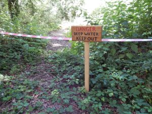 Wooden warning sign reading "DANGER DEEP WATER KEEP OUT" on a post, with red and white hazard tape strung across a woodland path.